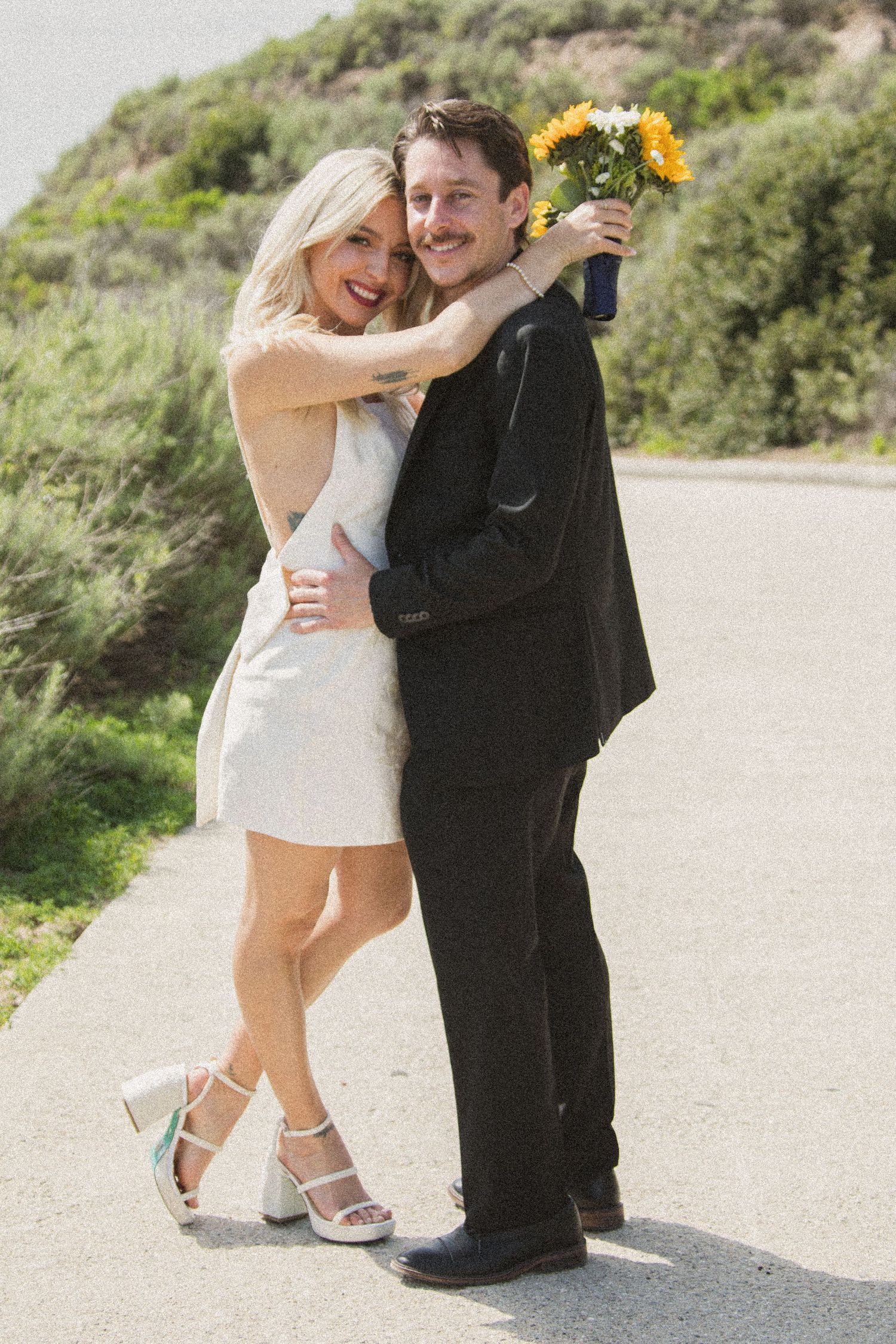 Playful wedding couple poses together on a coastal path with sunflowers and natural scenery in the background.