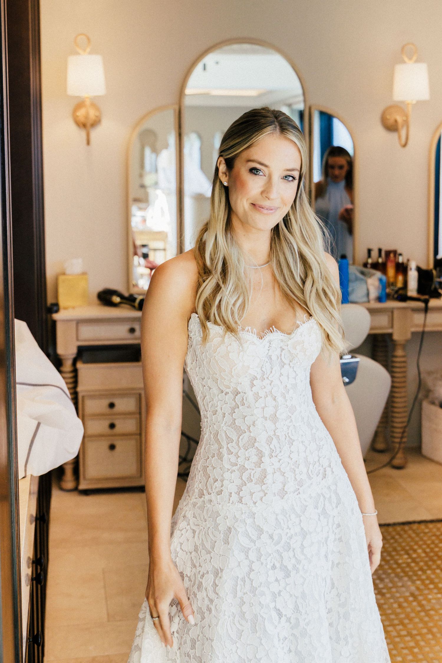 Bride wearing a white lace wedding dress poses in an elegant room with warm lighting and wall sconces.