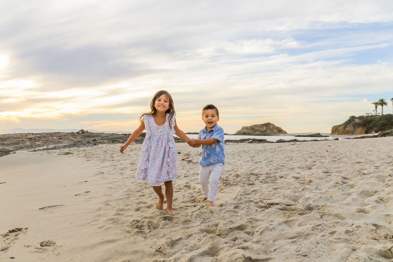 Two children run joyfully along the beach at sunset with rocky coastline in the background.