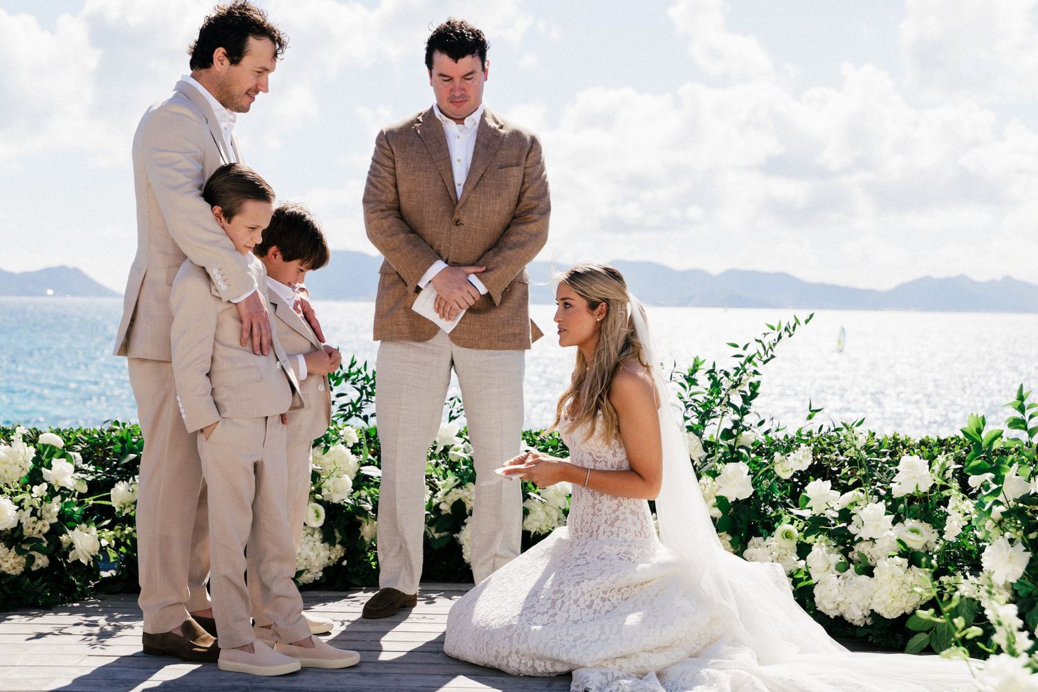 Emotional wedding ceremony moment captured against a stunning ocean backdrop with white floral decor.