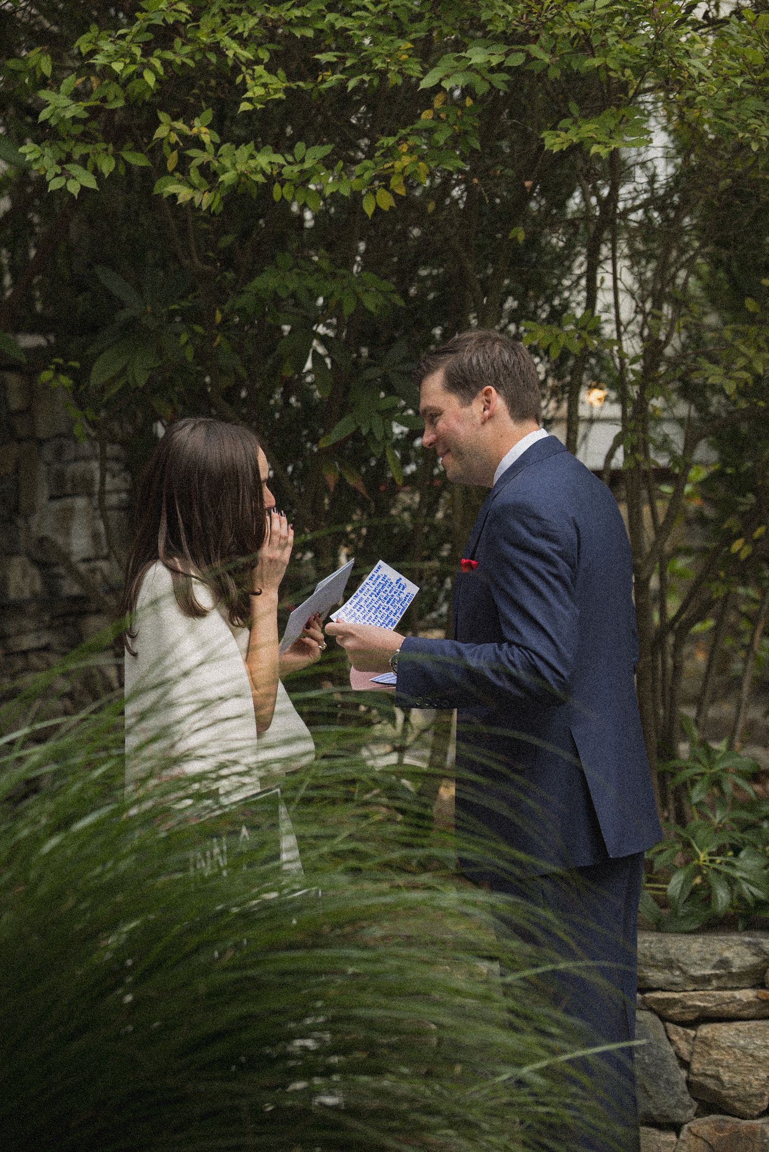 A smiling groom in a navy suit reads a blue card aloud while his emotional bride wipes her eye in a leafy garden.