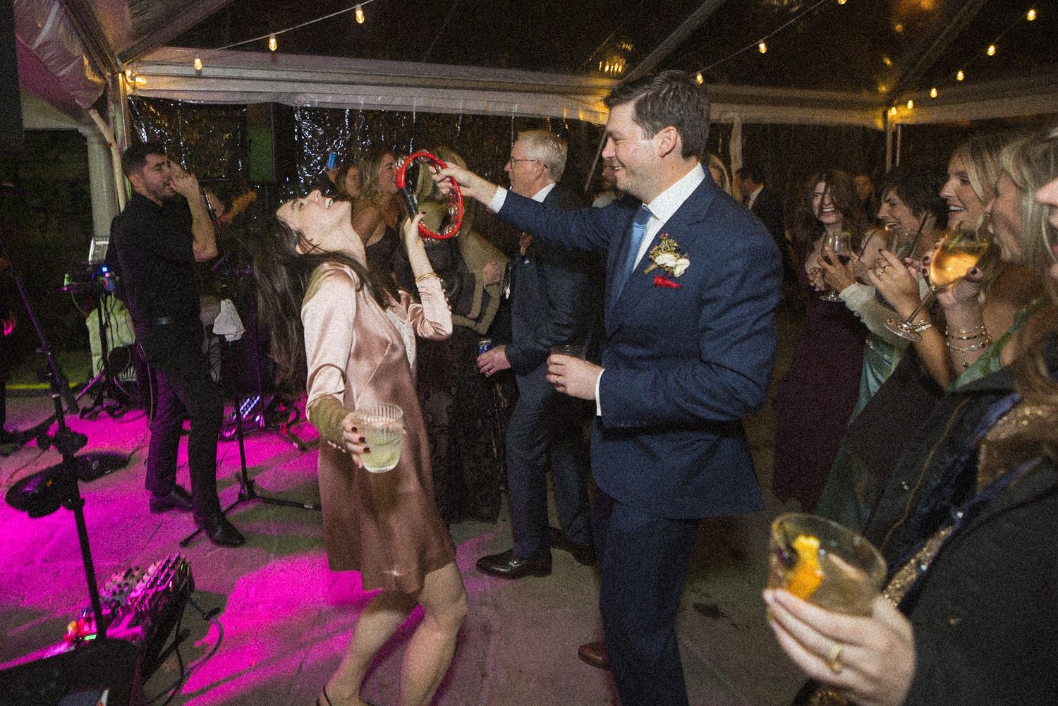 A groom in a navy suit dances with a woman in a blush dress as wedding guests with cocktails cheer under string lights.