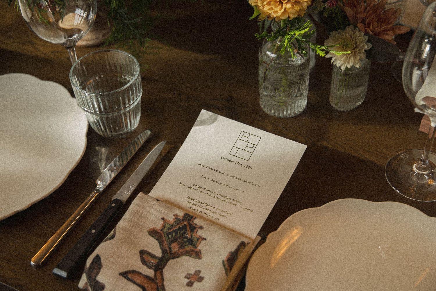 Close-up of a wedding place setting with a printed menu, folded patterned napkin, cutlery, glasses and small flower vases.