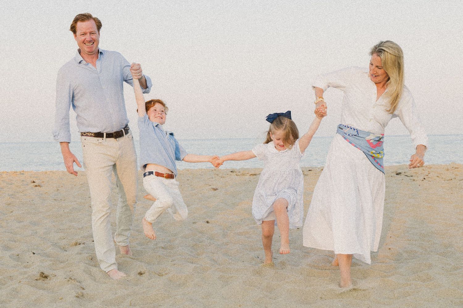 Family of four playing on a sandy beach at sunset, parents swinging children by their hands.