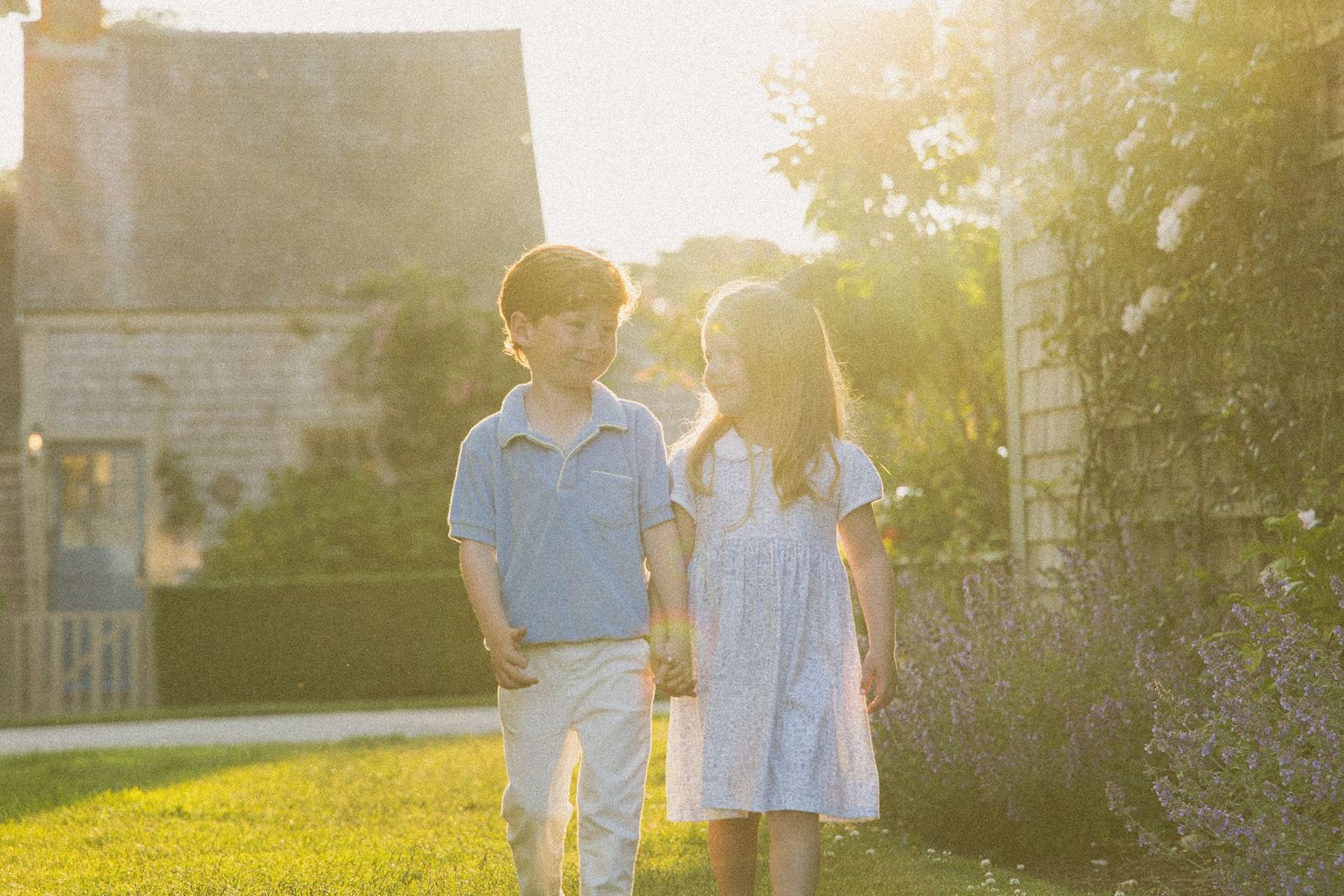 Two children walk hand in hand in a sunlit garden near a stone house during golden hour.