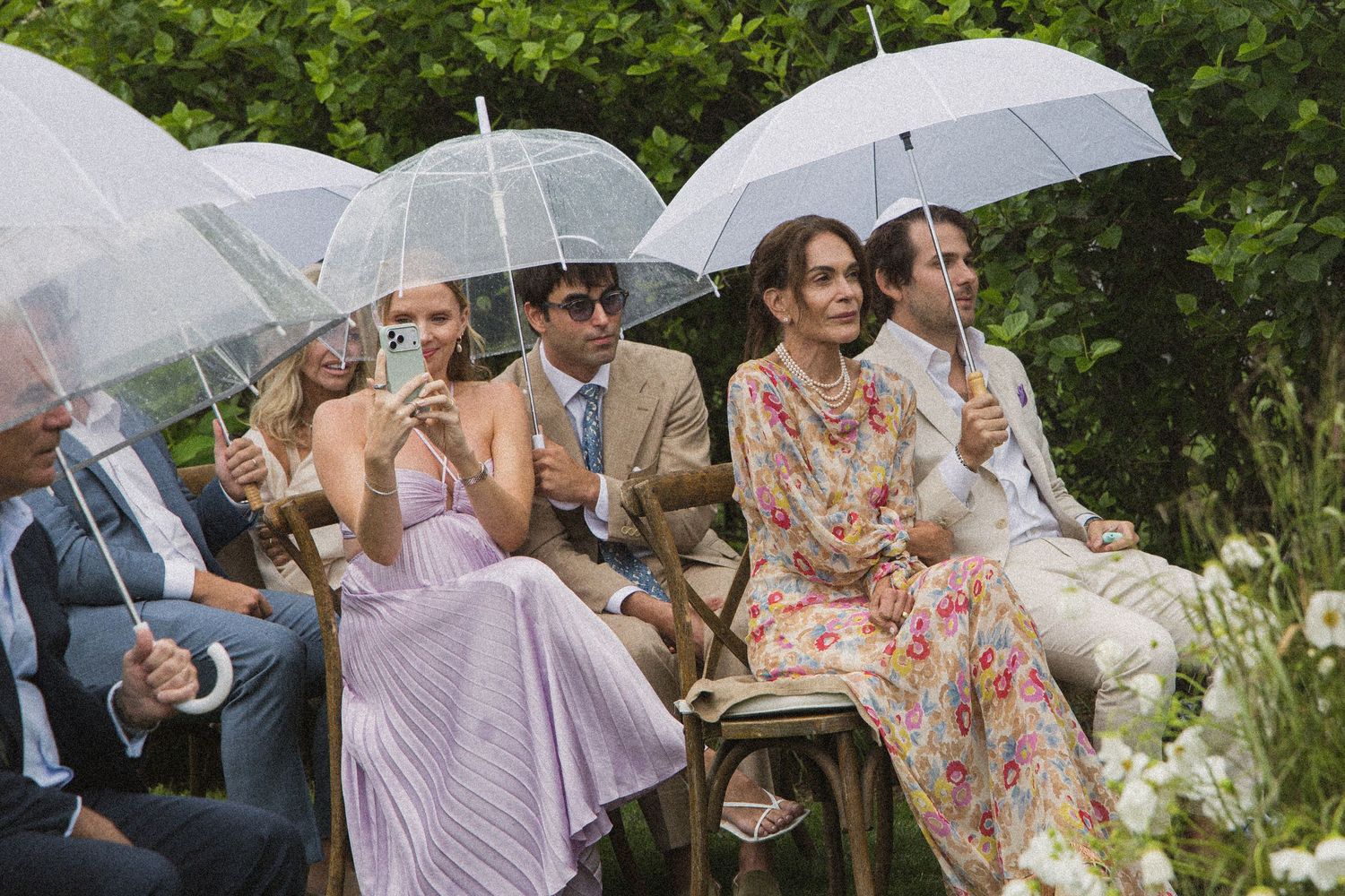 Wedding guests sitting outdoors holding clear umbrellas in elegant attire during an outdoor ceremony.
