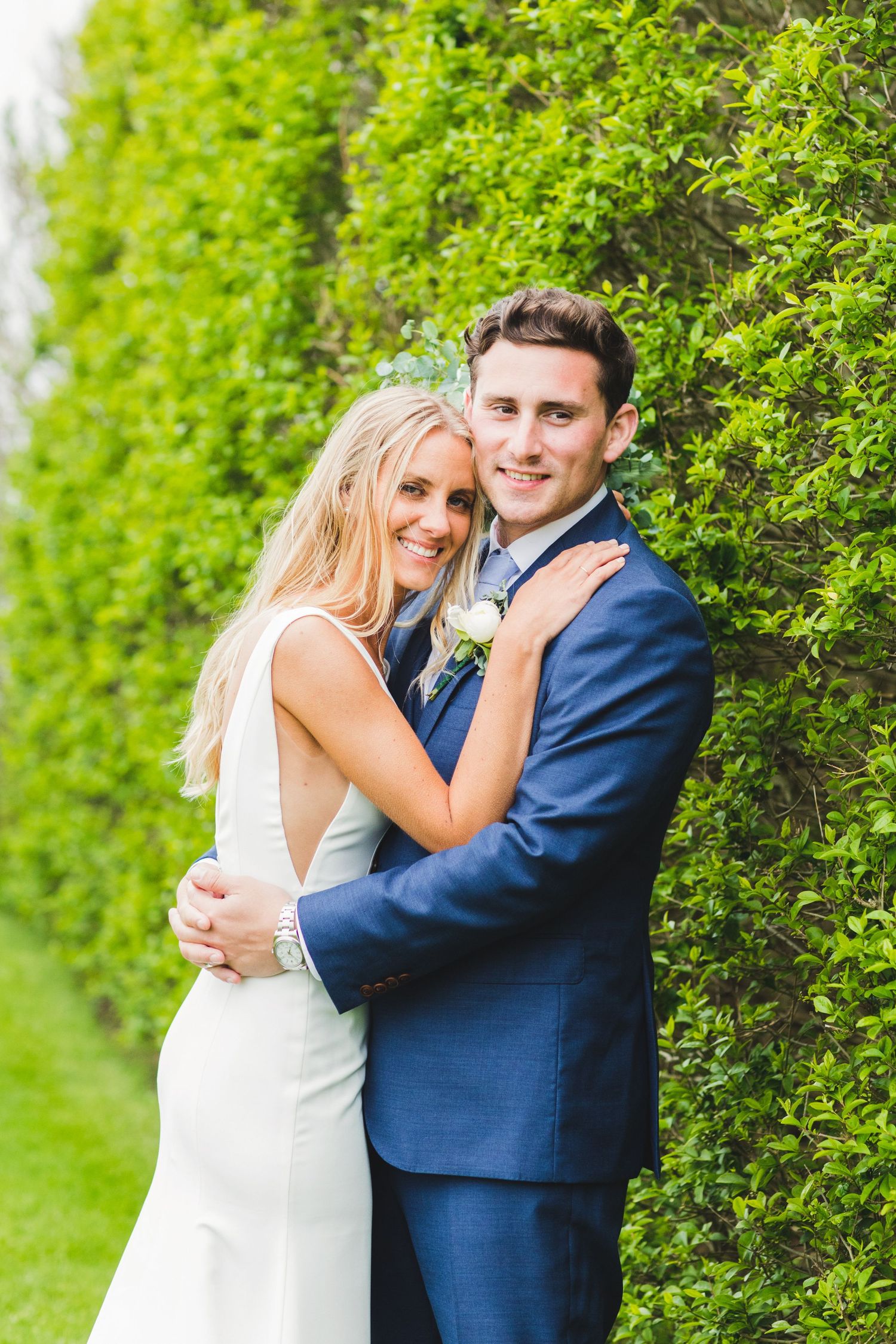 A couple in wedding attire embrace against a lush green hedge backdrop.