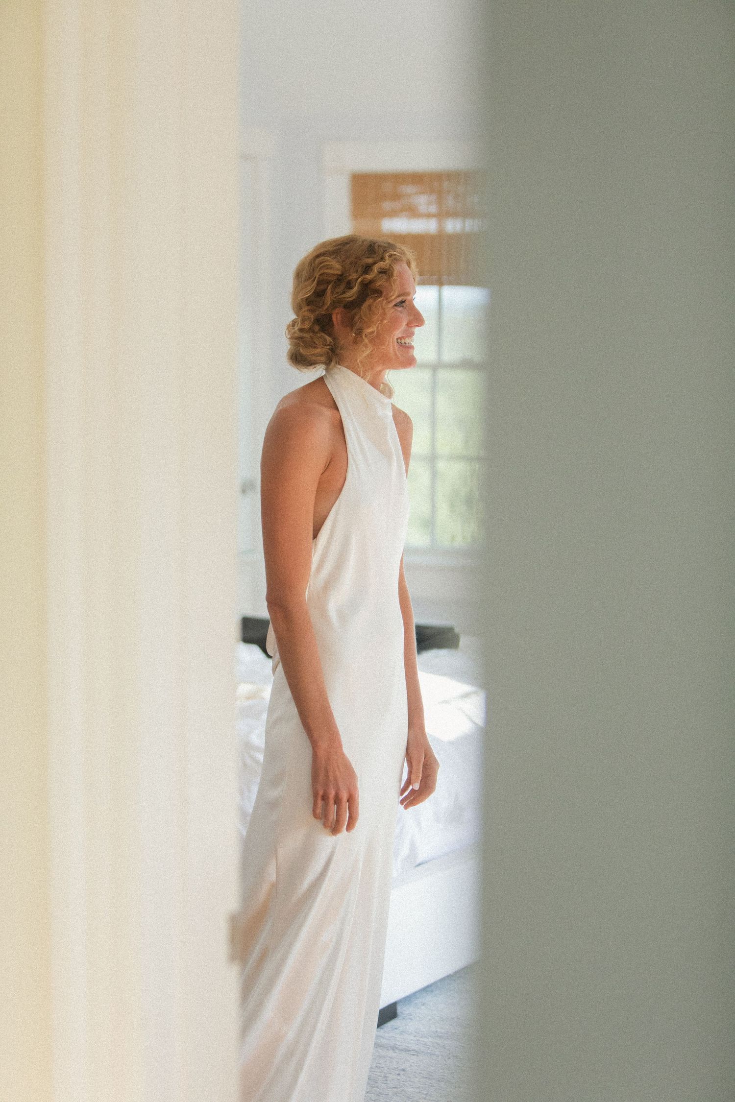 A person in an elegant white sleeveless dress stands in profile in a sunlit room.