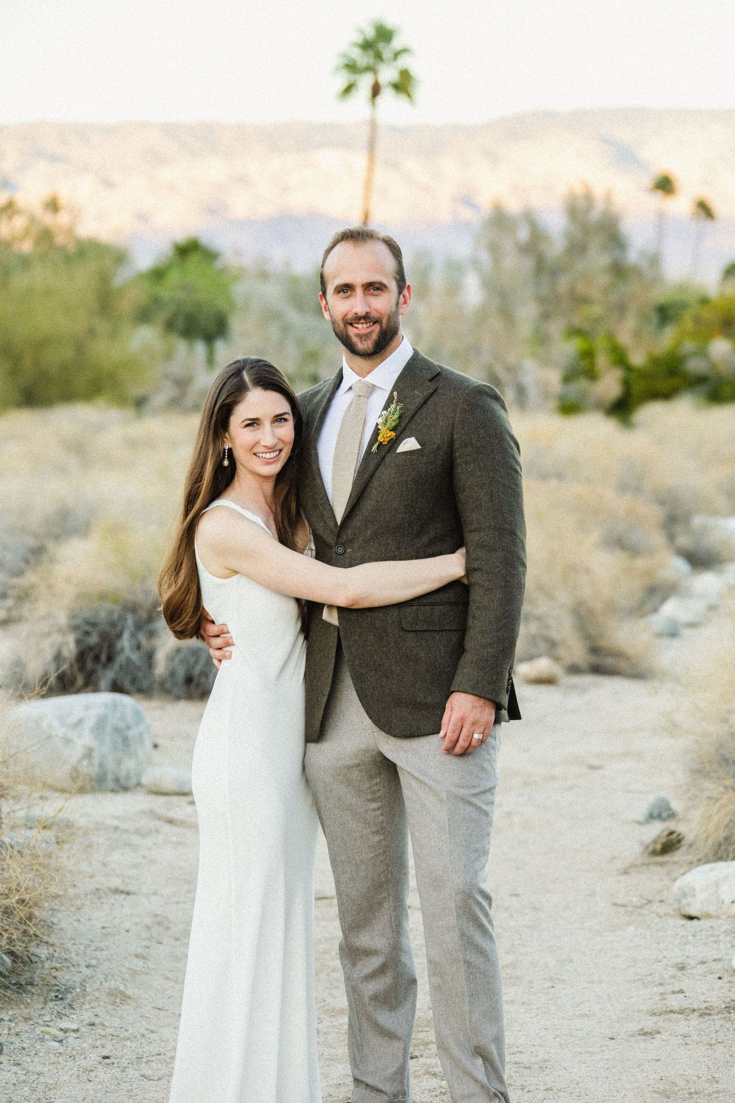 A couple poses for wedding photos in a desert landscape with mountains and palm trees.
