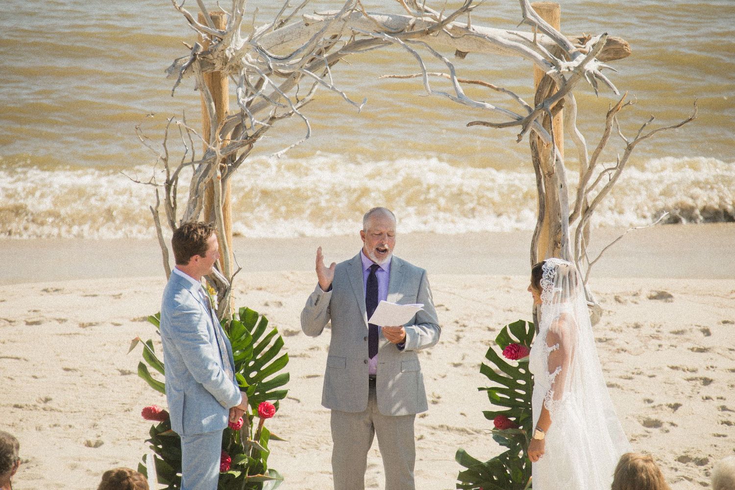 Beach wedding ceremony setup with driftwood arch and tropical flowers against ocean backdrop.