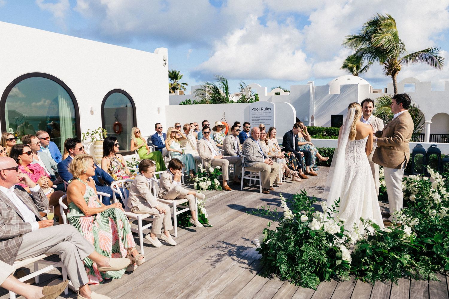 Wedding guests seated in white chairs on a beachfront deck overlooking turquoise waters.