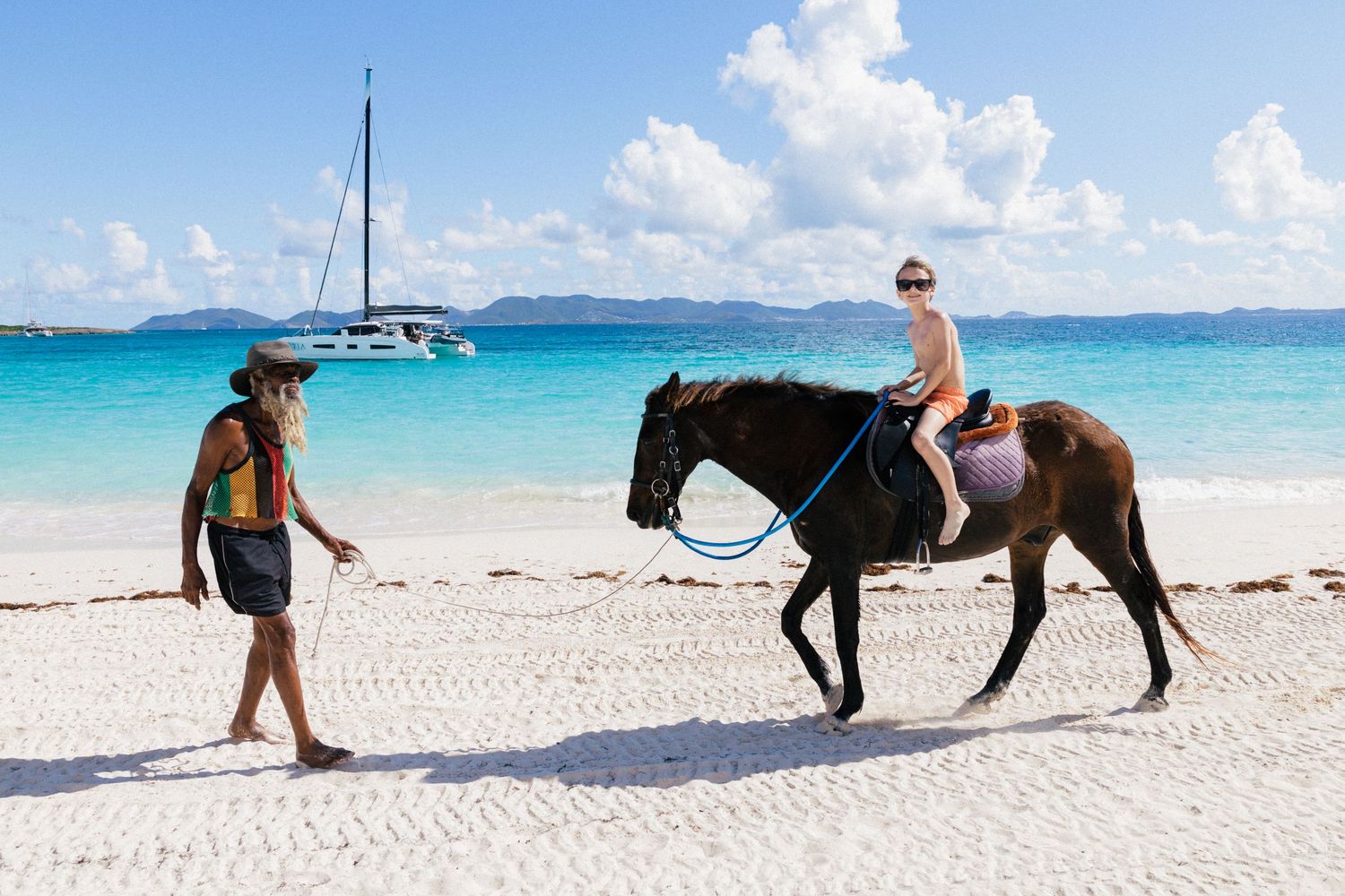 Couple in beachwear standing on white sand beach with turquoise ocean and mountains behind.