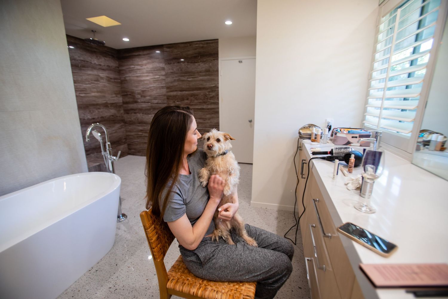 Person holding a small dog while sitting in a bathroom setting.