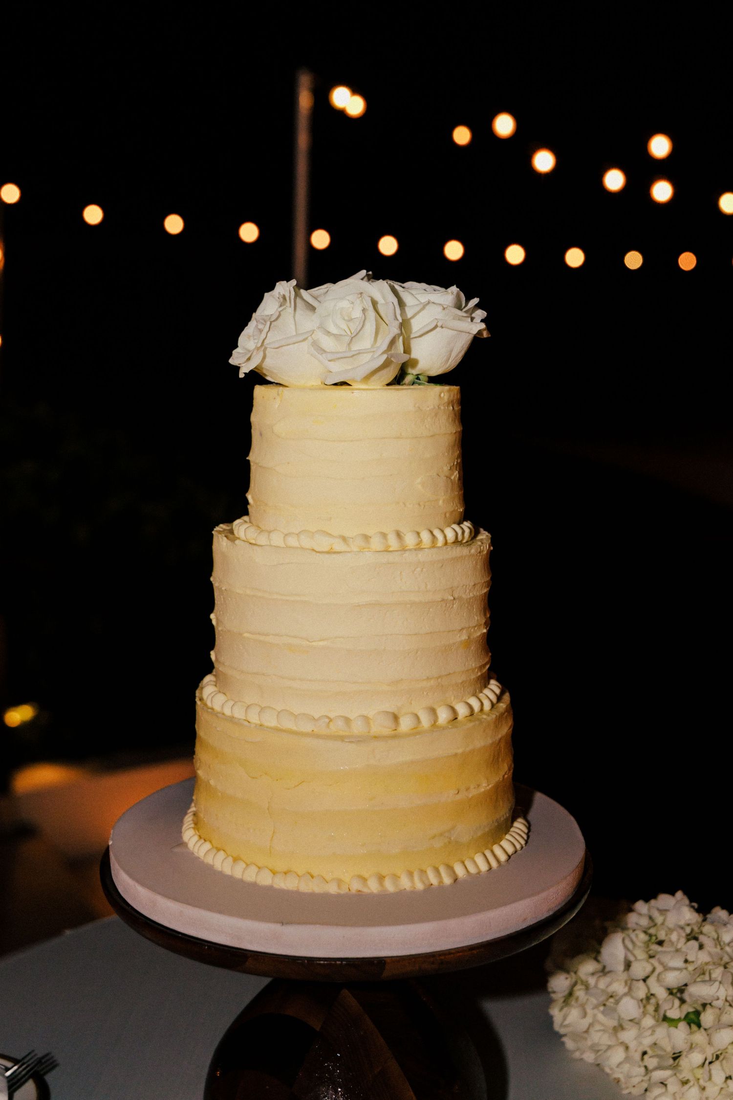 Three-tiered cream colored wedding cake decorated with white roses on display at night.
