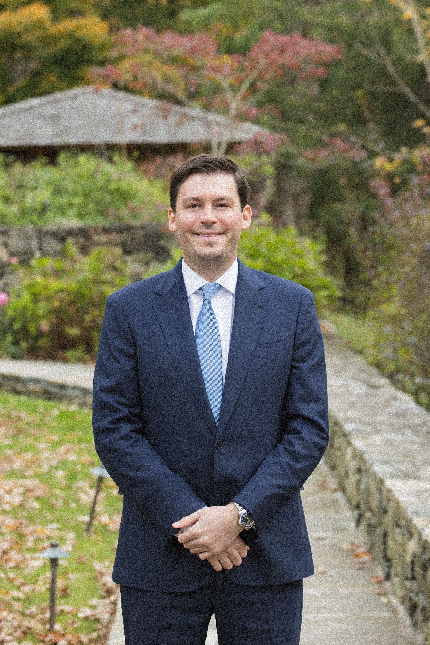 Smiling man in a navy suit and light blue tie standing outdoors in a lush garden with a stone wall and gazebo.