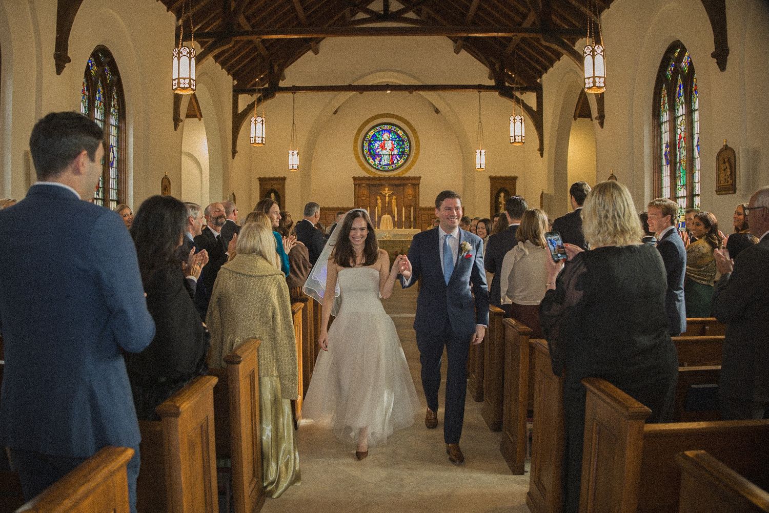 Bride and groom hold hands and walk down the aisle of a wooden church as smiling wedding guests applaud from the pews.