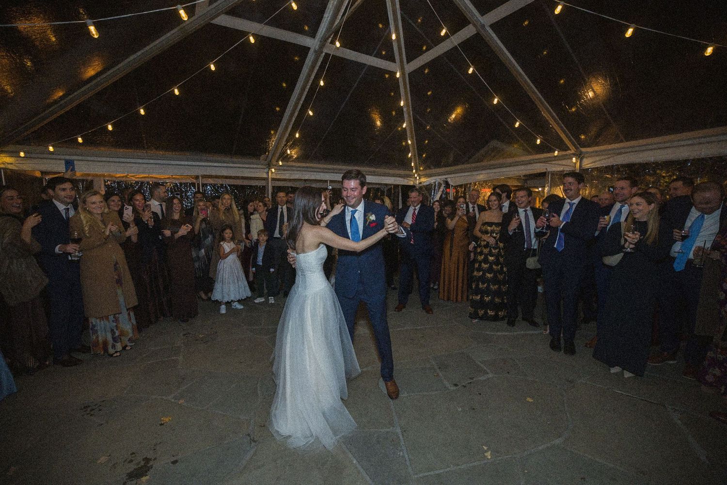 Bride in a white gown and groom in a dark suit share a first dance under a tented ceiling with string lights while wedding guests watch.