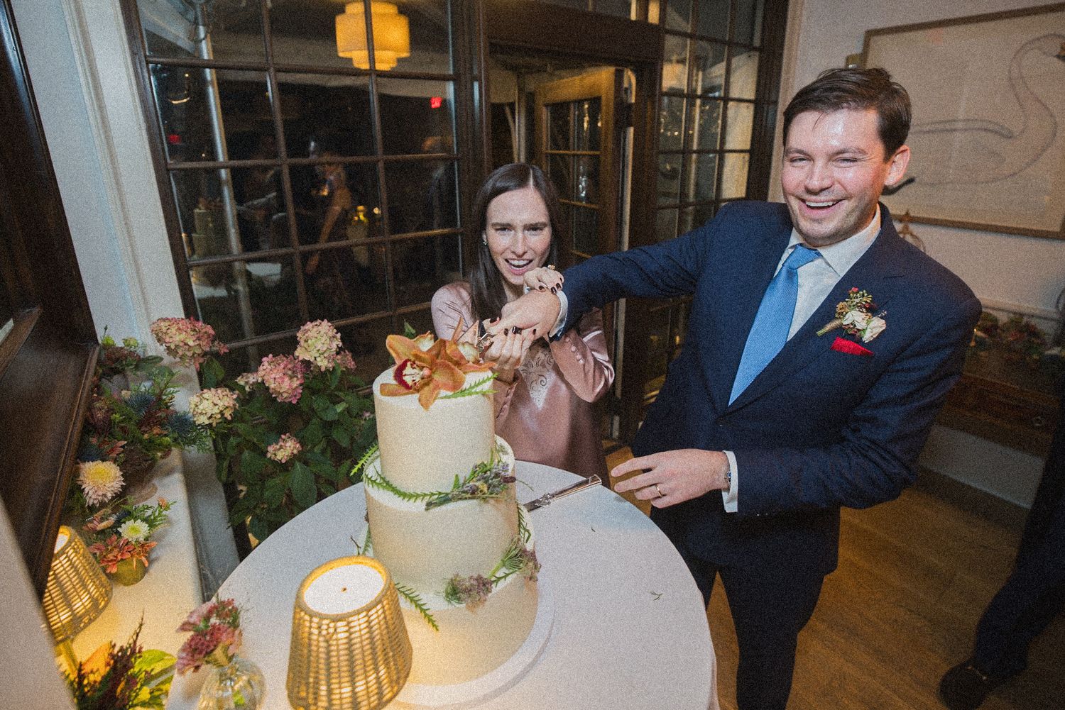 A smiling couple stands together and cuts a tiered wedding cake decorated with fresh flowers and greenery.