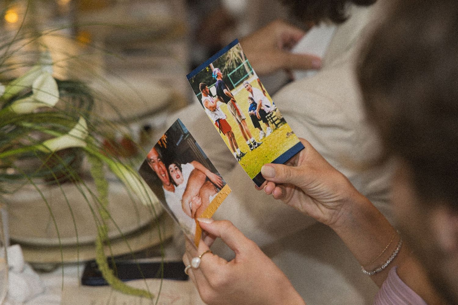 Hands holding printed photos at a gathering, with a floral centerpiece visible in the background.