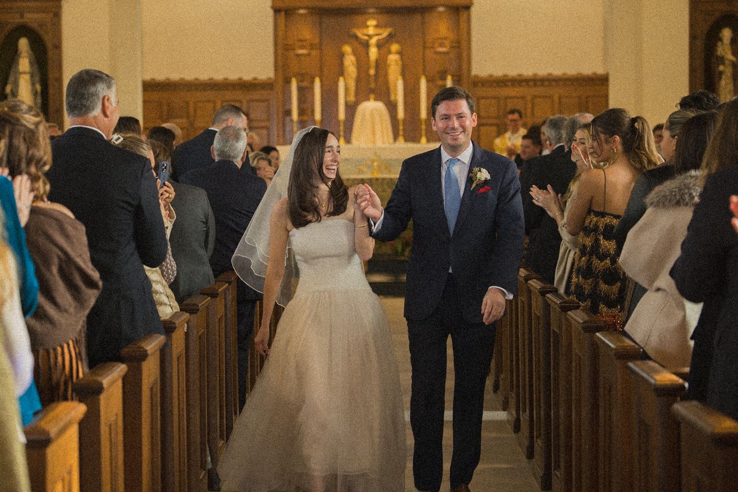 Bride in a white gown and groom in a navy suit hold hands and walk down the church aisle past smiling wedding guests.