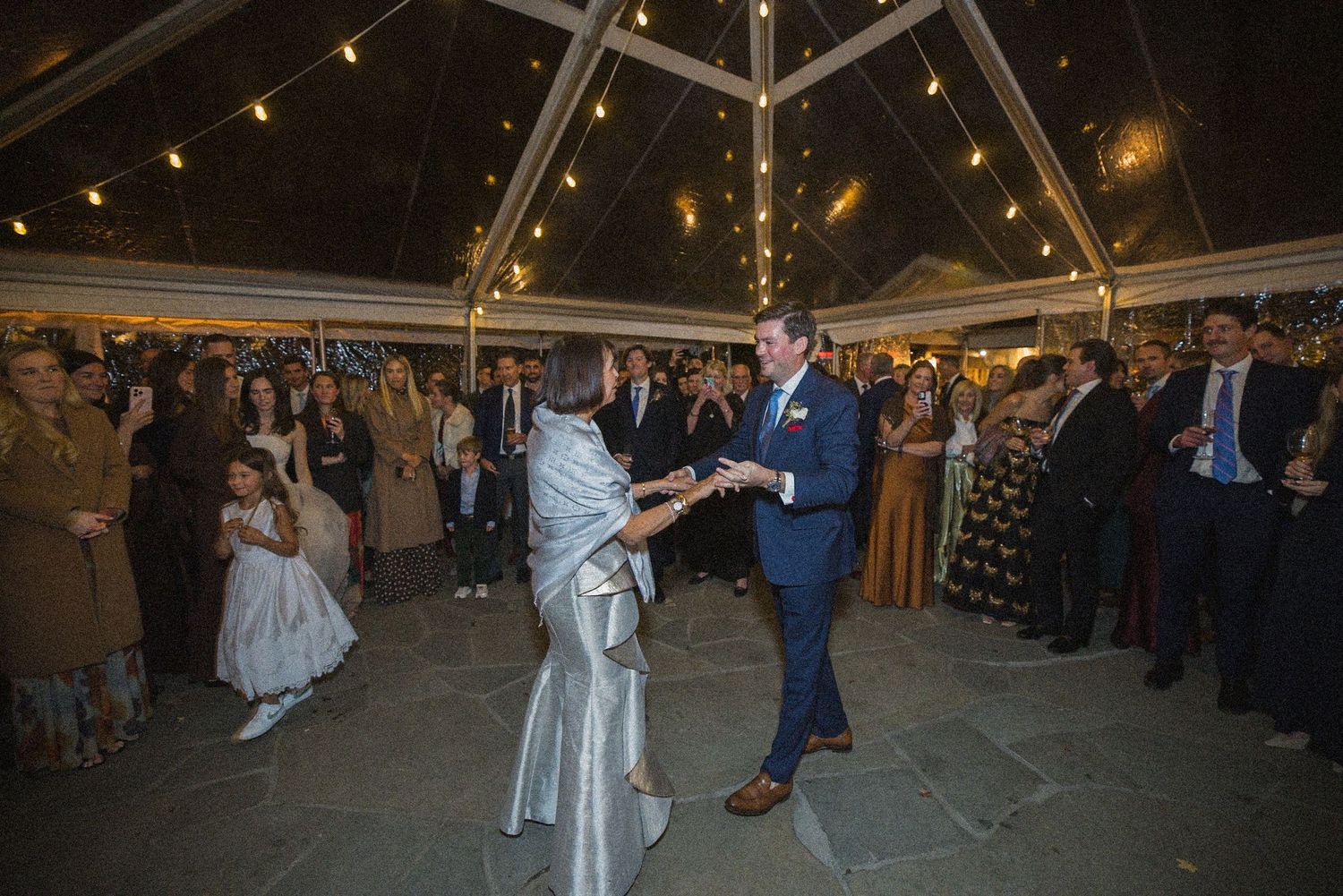 Bride in a silver gown and groom in a navy suit share a first dance under a tented wedding reception as guests watch.