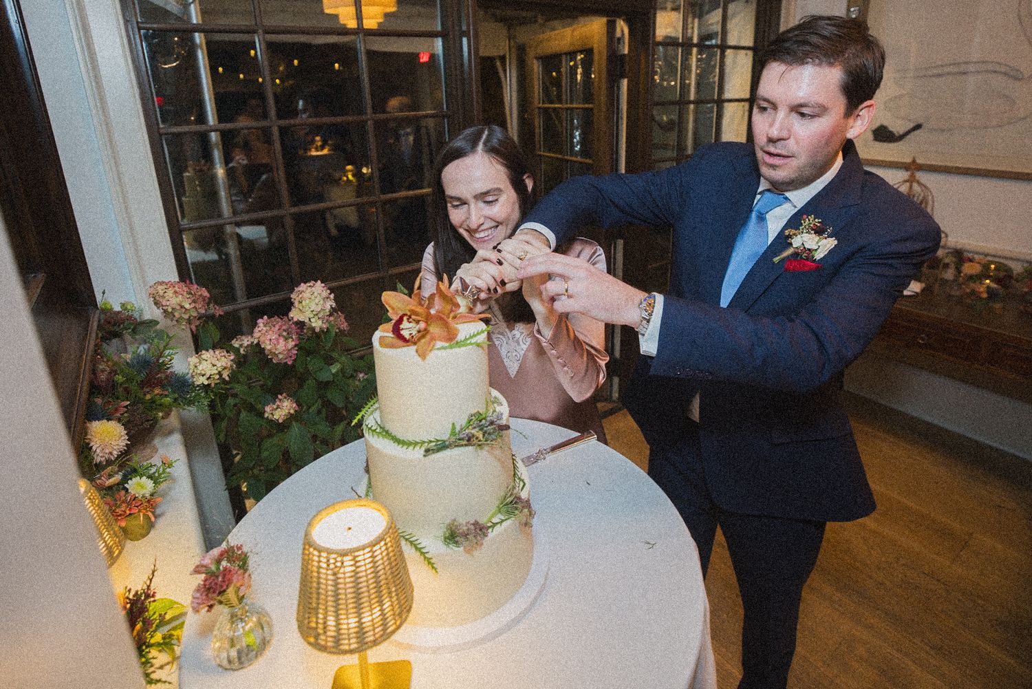 A bride and groom in a navy suit cut their floral-decorated tiered wedding cake together at a candlelit reception.