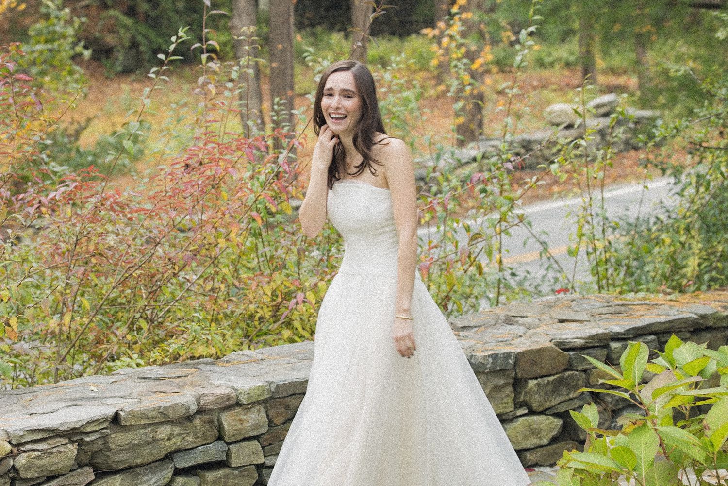 Smiling bride in a strapless white wedding gown standing beside a stone wall surrounded by colorful autumn foliage.