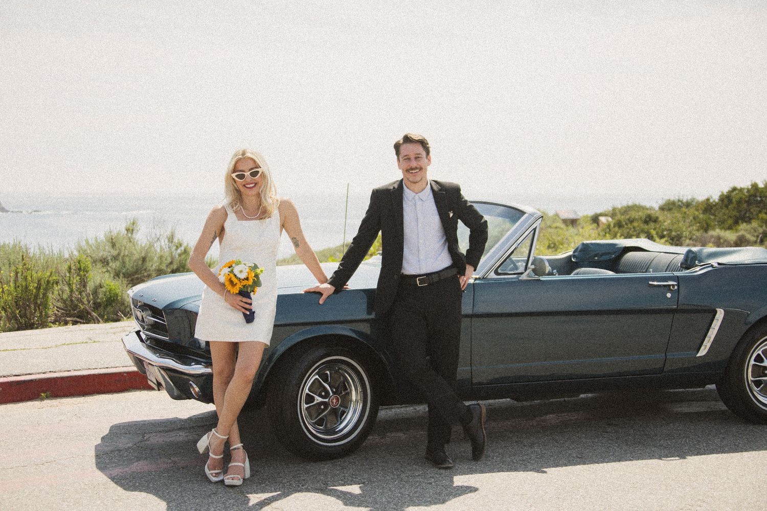 Stylish couple in formal attire posing with classic Ford Mustang on mountain road.