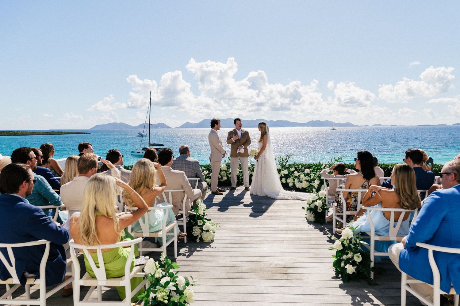 Oceanfront wedding ceremony setup with white chairs and floral decorations along a wooden boardwalk.
