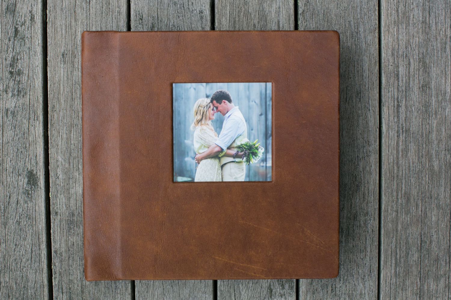 Leather-bound wedding album with small photo insets displayed on rustic wooden planks.