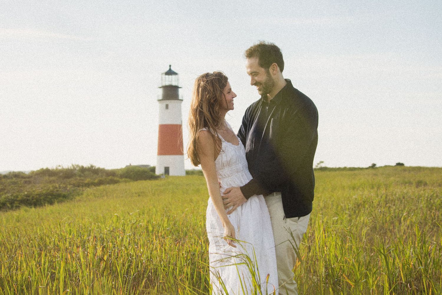 A couple embraces in a grassy field with a lighthouse visible in the background during golden hour.
