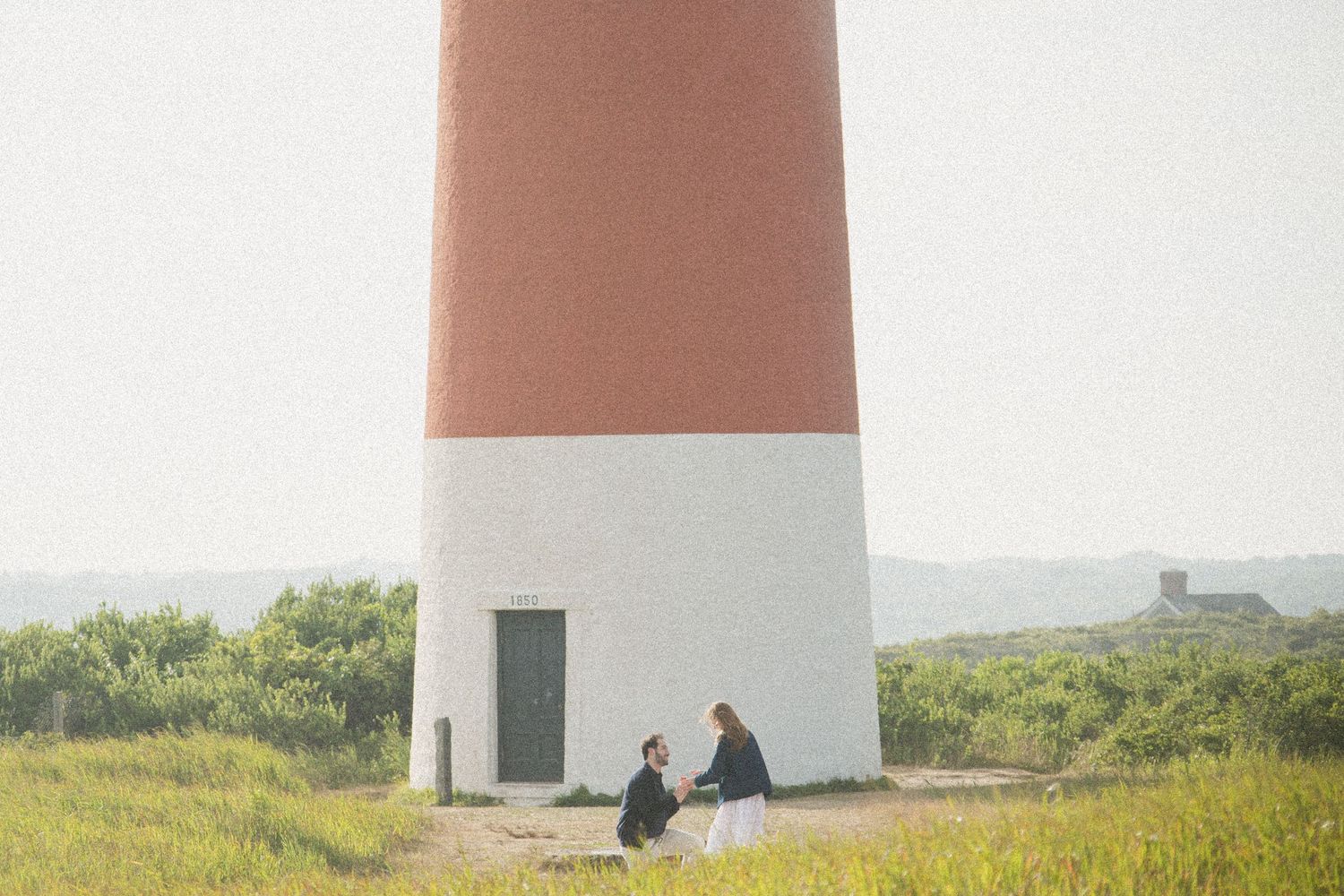Two people sitting at the base of a red and white lighthouse surrounded by green grass.
