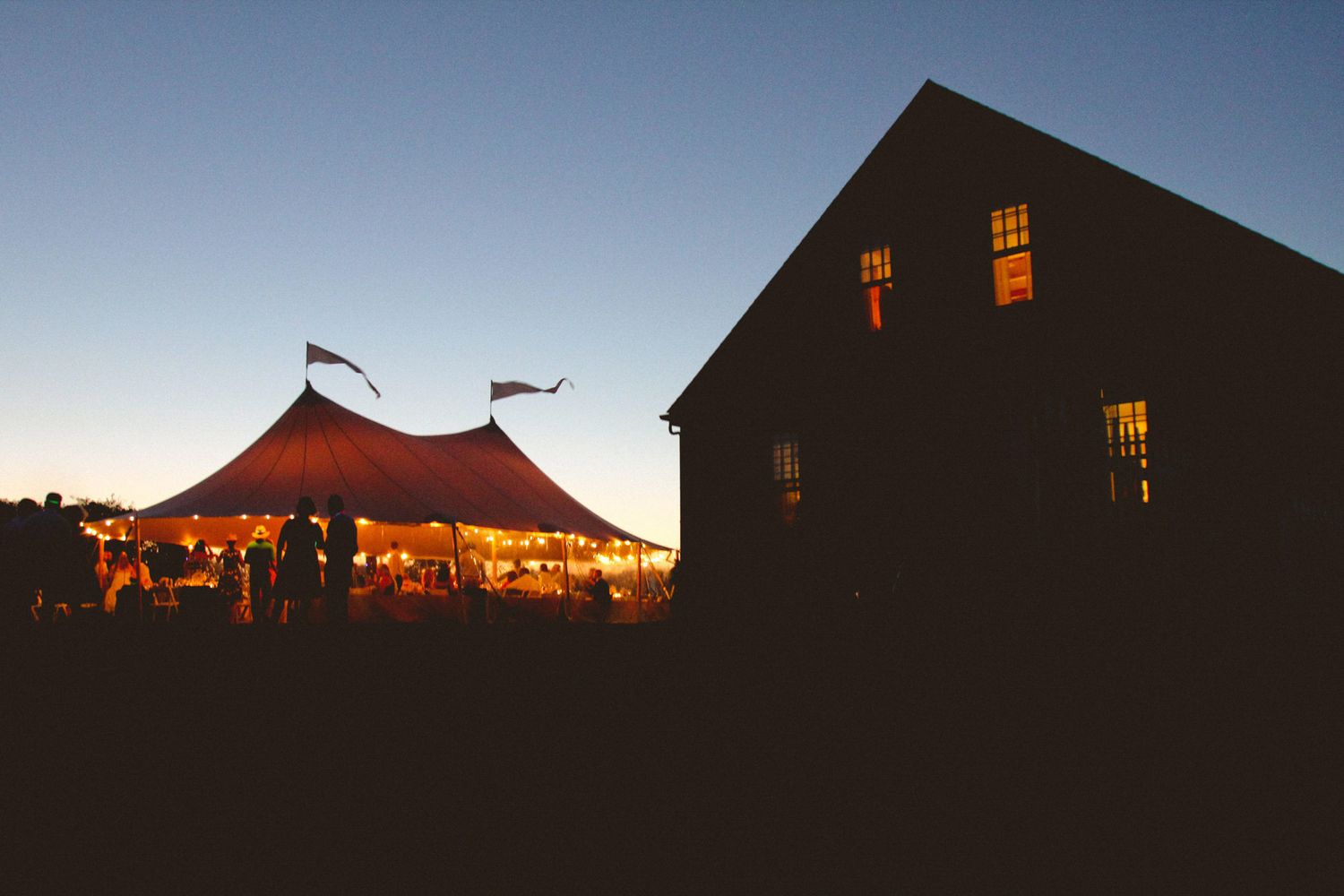 Glowing wedding tent with flags at dusk beside a dark silhouetted barn building at twilight.