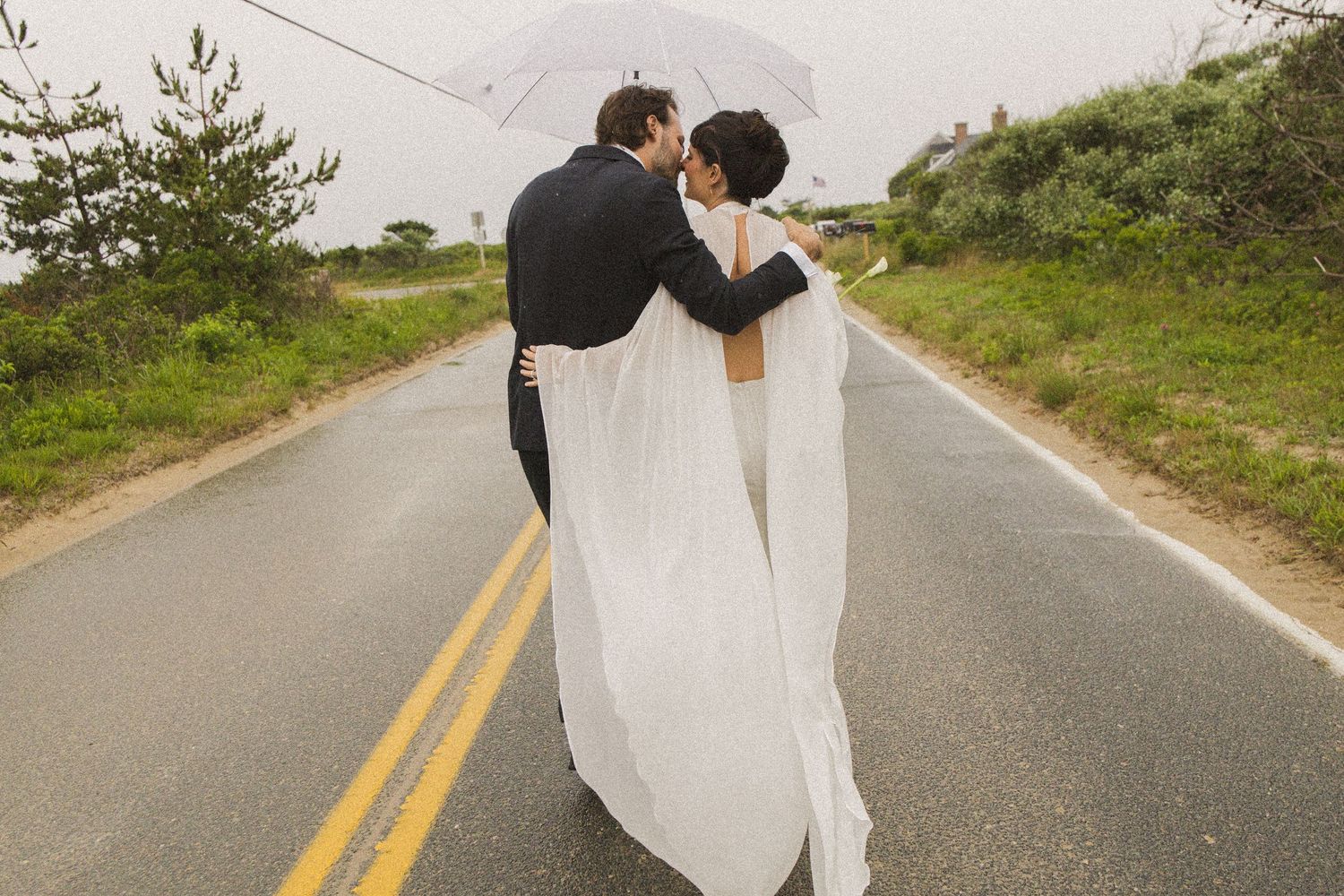 Newlywed couple walking down a scenic coastal road, bride in flowing white gown, groom in black tuxedo, sharing a kiss.