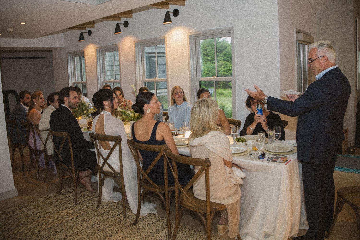 Elegant wedding rehearsal dinner with guests seated at a long table while a man in a suit gives a toast.