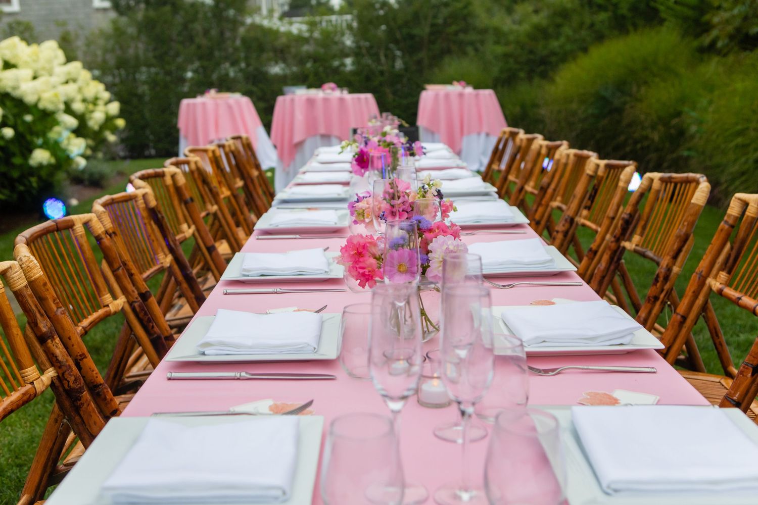 Elegant outdoor pink tablescape with floral centerpieces, white place settings, and bamboo chairs for a garden party.