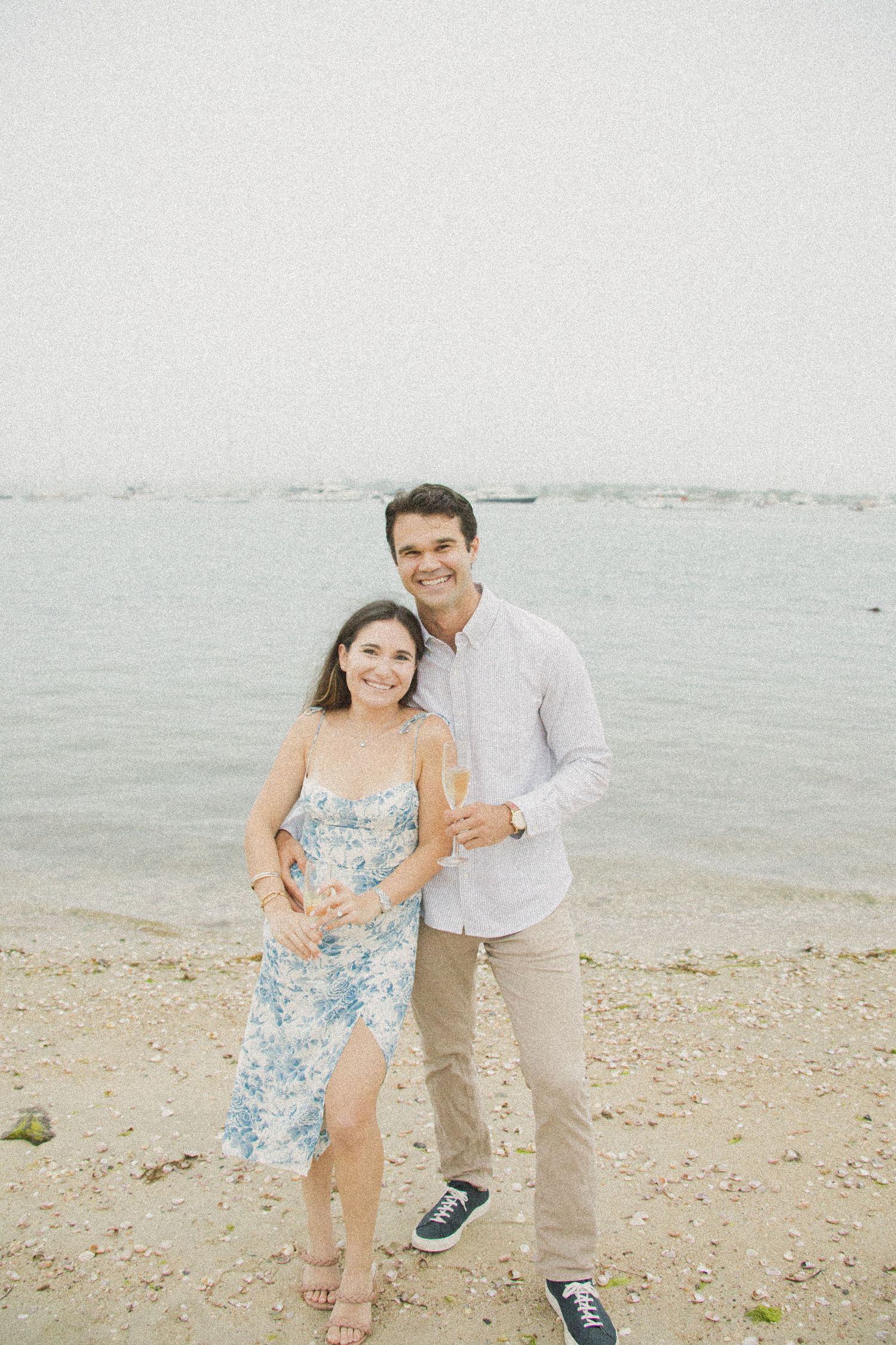 Couple smiling on sandy beach holding champagne glasses, woman in floral dress, man in light blue shirt, ocean backdrop.