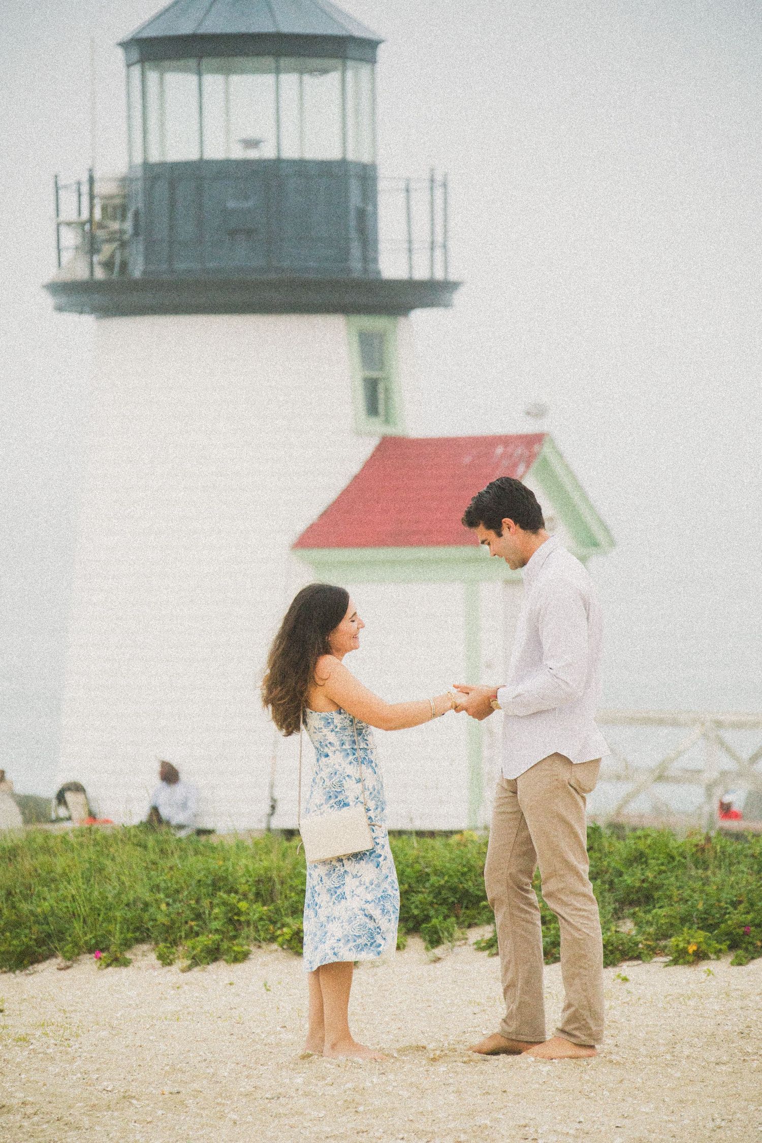 Couple holding hands on sandy beach in front of white lighthouse with red roof on Nantucket island.
