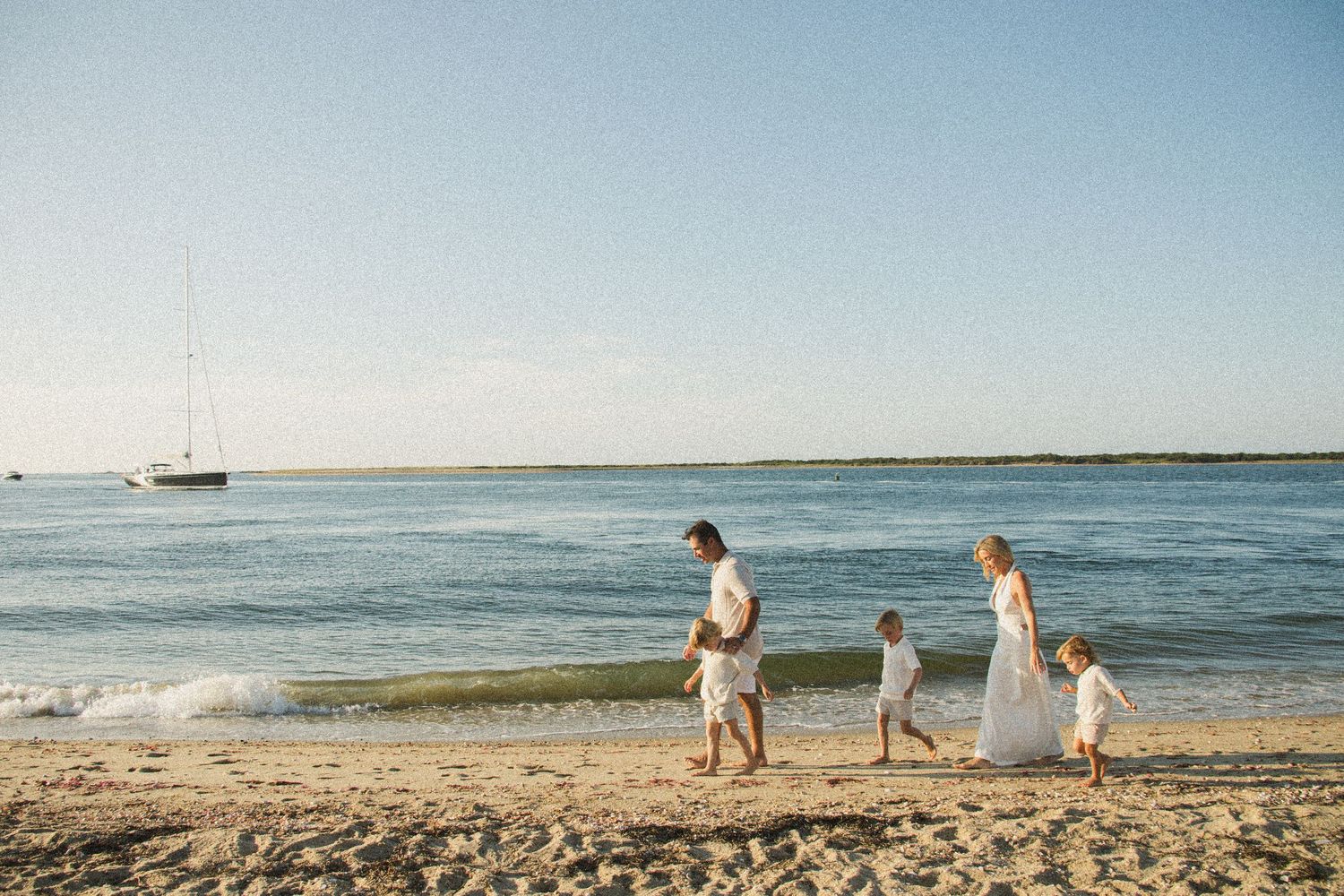 Family of four walking along a sandy beach shore in white outfits with a sailboat in the background.