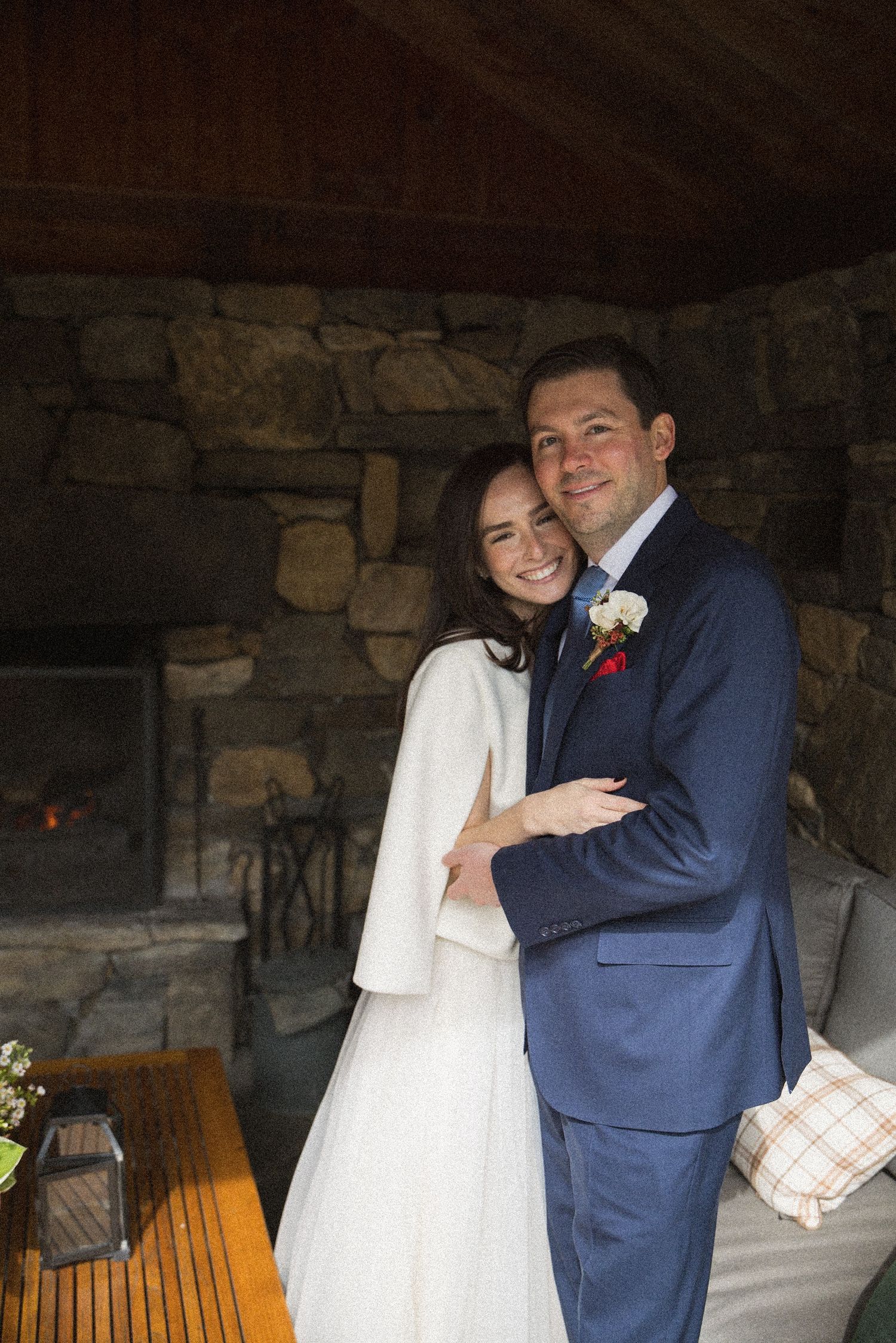 Smiling bride in a white dress and shawl embraces her groom in a navy suit beside a rustic stone fireplace at an intimate wedding.