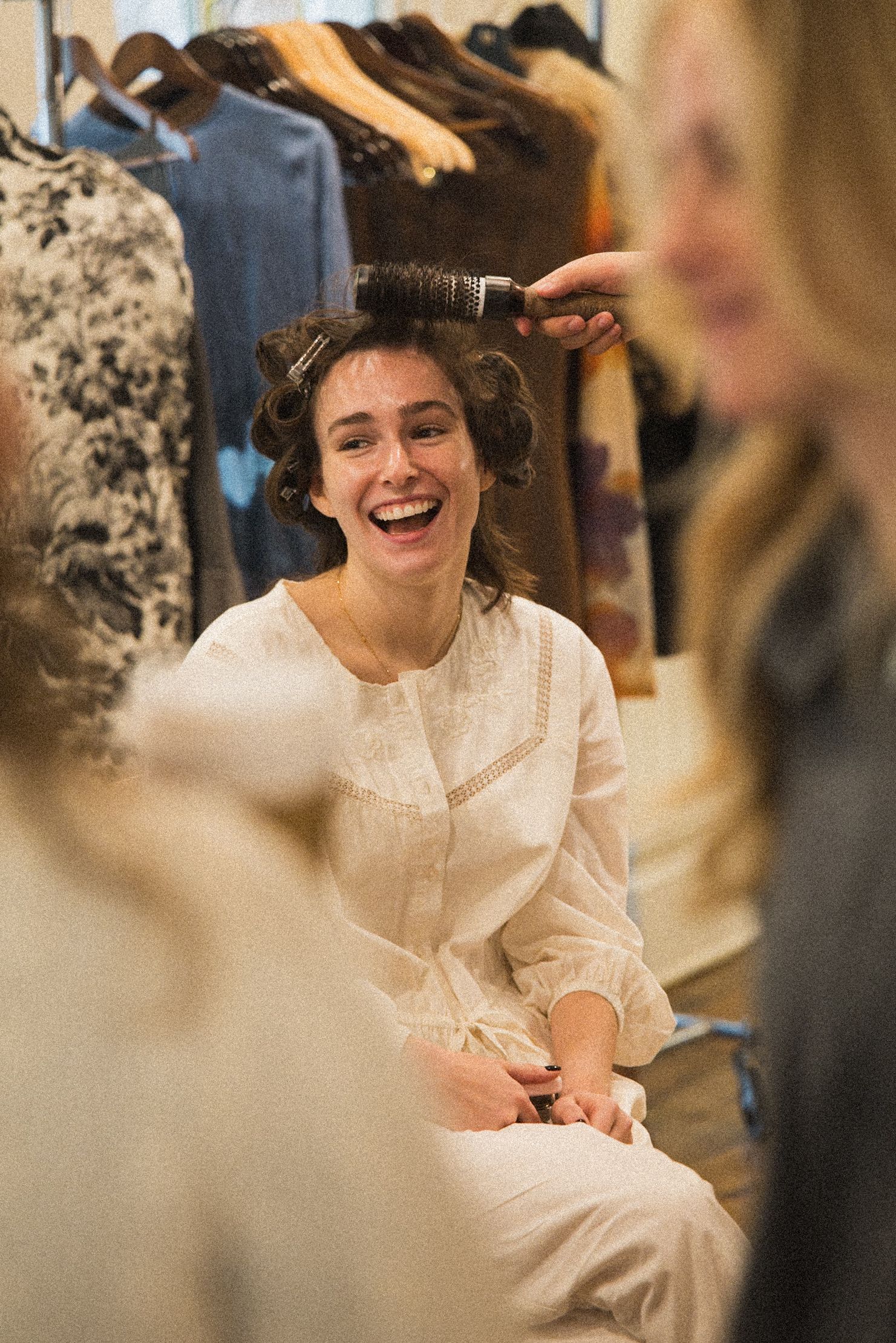 A smiling woman in a white blouse laughs while sitting with hair in curlers as a stylist brushes her hair beside racks of clothing.
