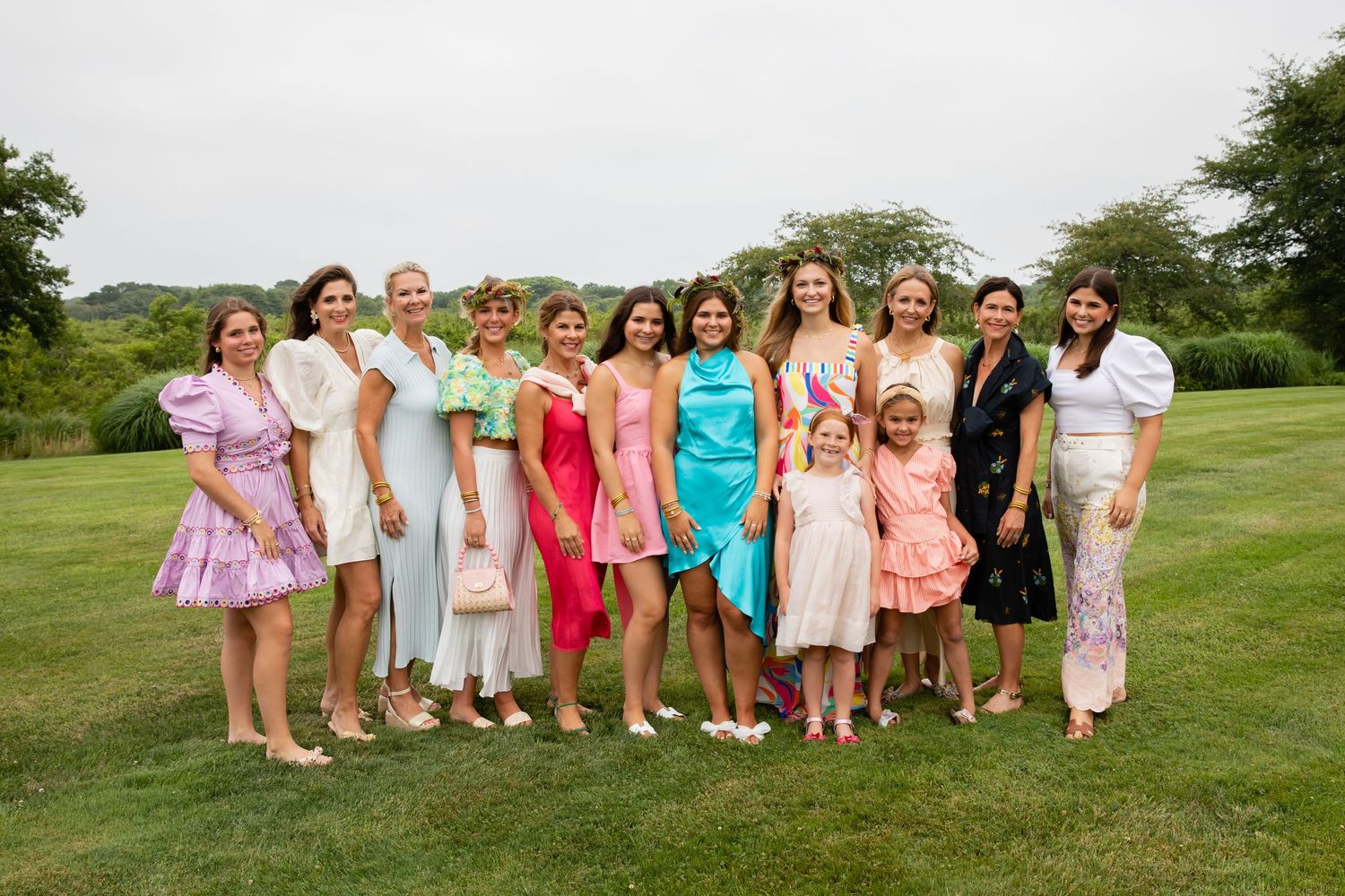 Group of women and girls in colorful dresses posing together on a green lawn at an outdoor celebration.