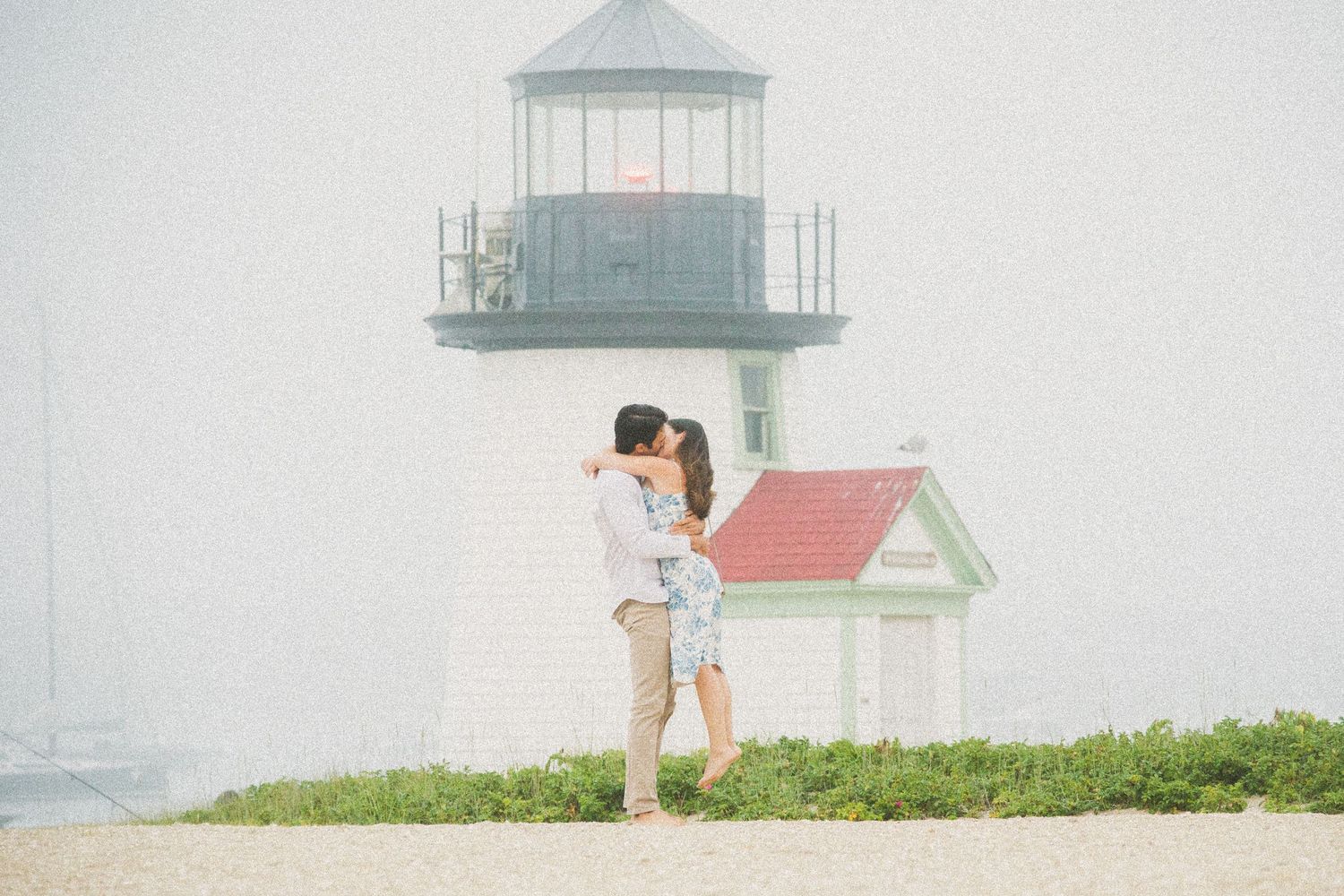 Couple embracing near a white lighthouse on a misty beach, romantic engagement photo with soft dreamy tones.