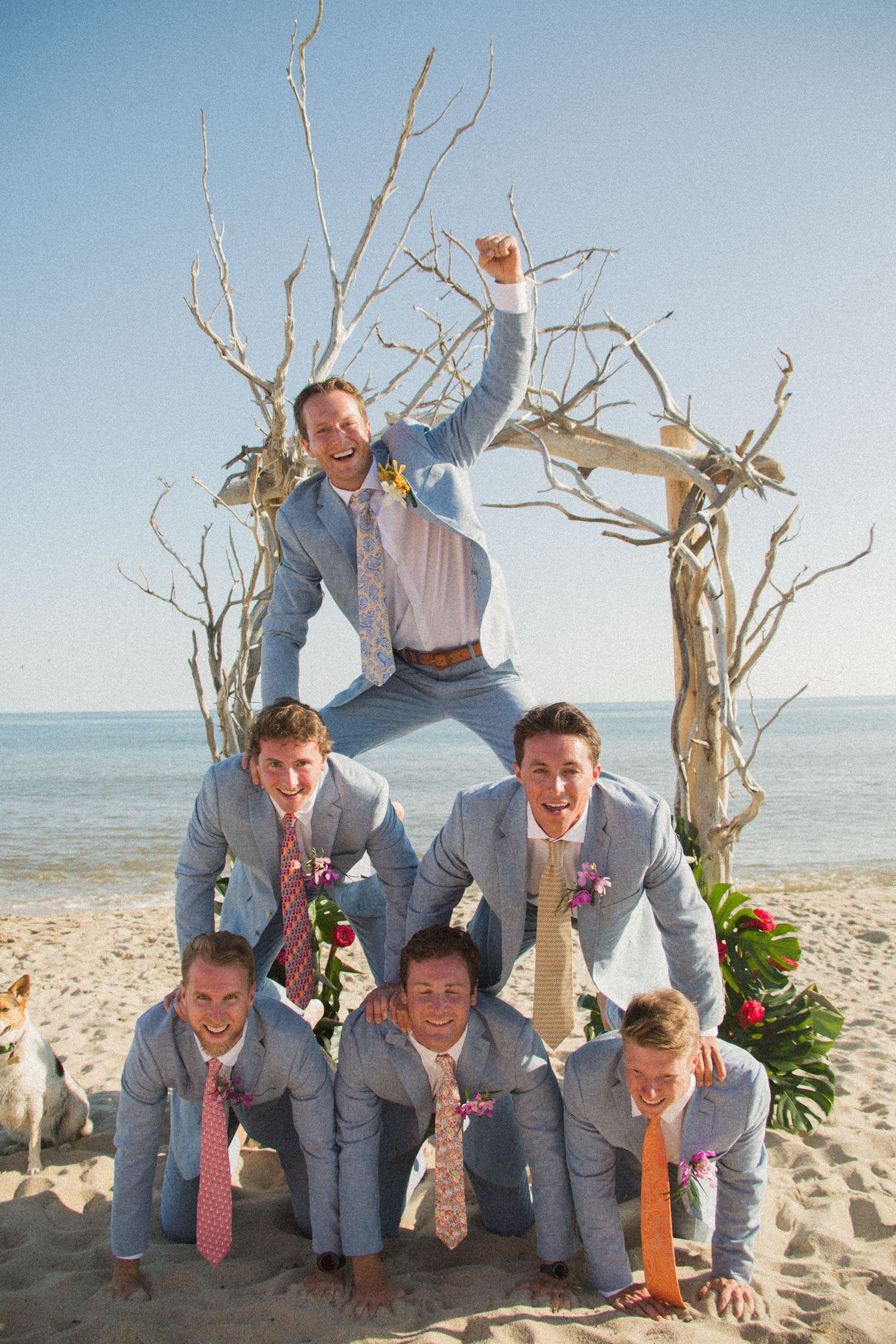Groomsmen in light blue suits form human pyramid on beach with driftwood arch.