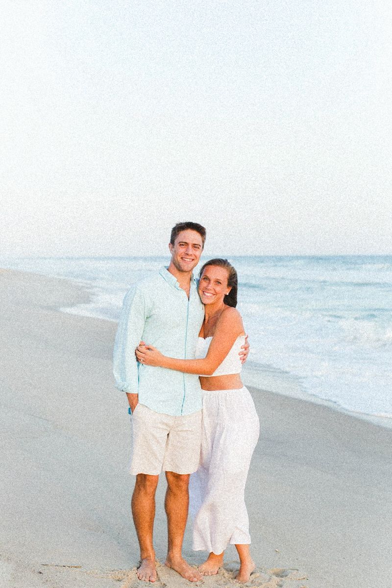 Couple walking along a serene beach at sunset in coordinated white and light blue outfits.
