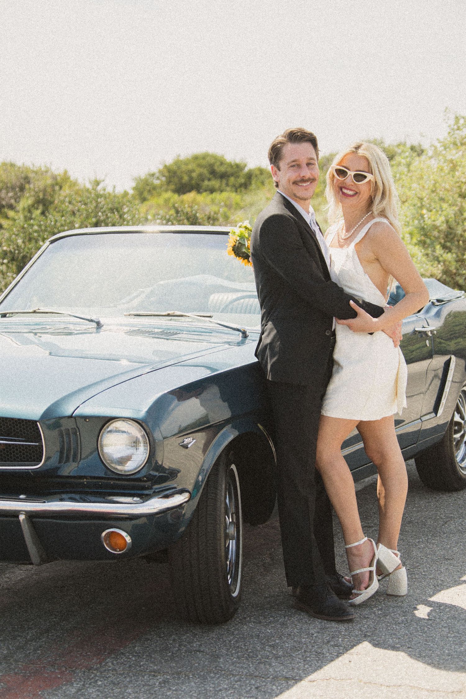 Playful couple poses by vintage Ford Mustang during outdoor photoshoot.
