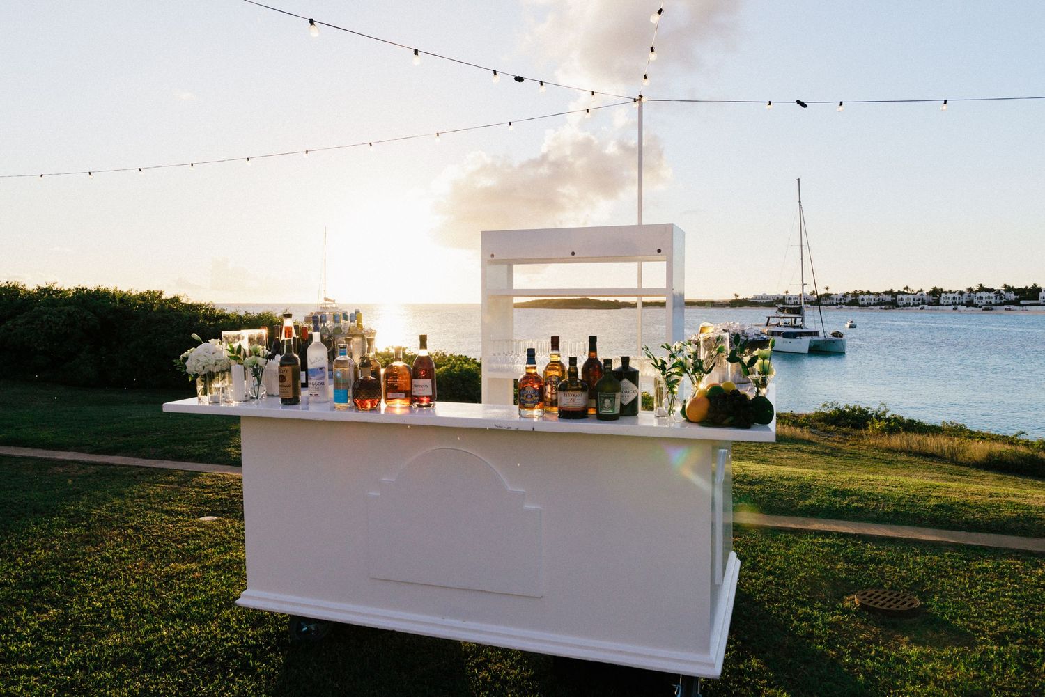 White outdoor bar setup with string lights and waterfront view at sunset.