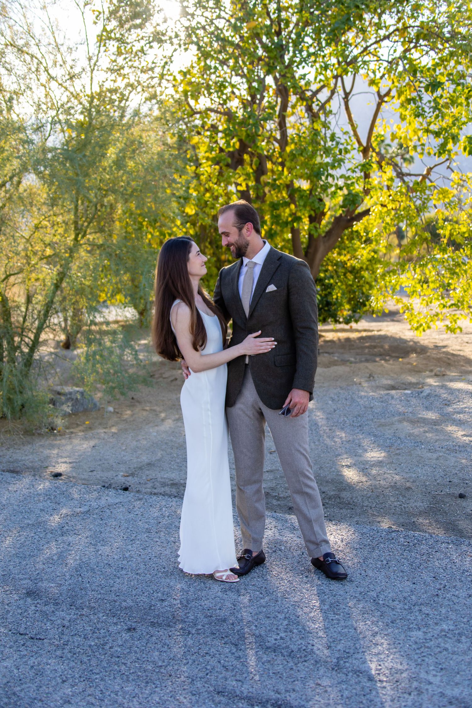 Couple in wedding attire embracing on desert path with trees.