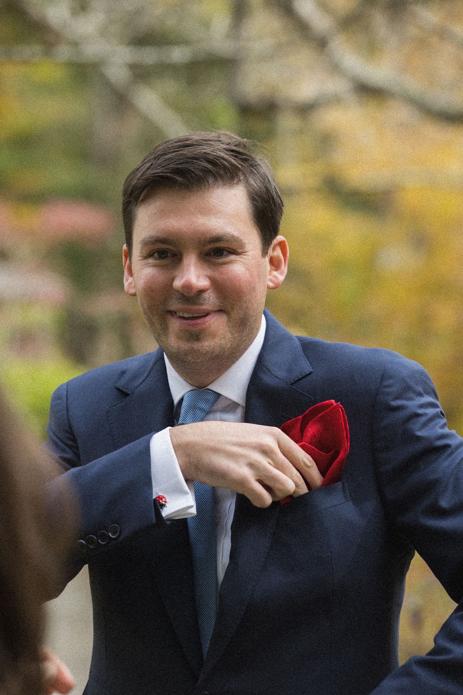 Smiling man in a navy suit and blue tie adjusts a red pocket square outdoors, with colorful autumn foliage blurred in the background.