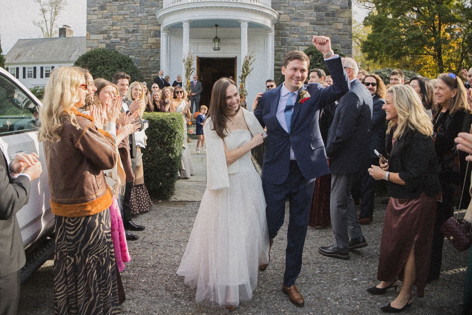 Smiling bride in a white dress and groom in a navy suit raise a fist as wedding guests cheer along a gravel path outside a stone building.